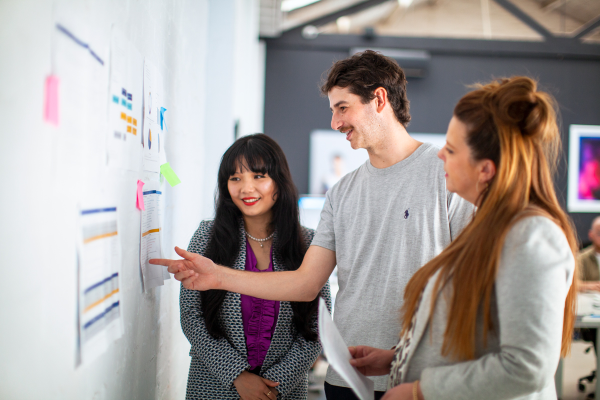2 females and a male pointing at a paper in a wall